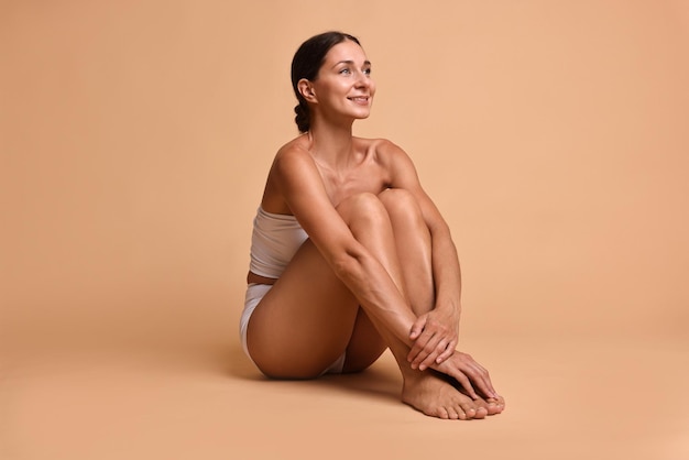 Donna sorridente con la pelle perfetta seduta su uno sfondo beige cura del corpo