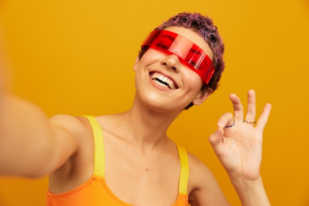 Donna sorridente con occhiali insoliti millenaria che scatta selfie in abbigliamento sportivo su uno spazio libero sullo sfondo di uno studio arancione