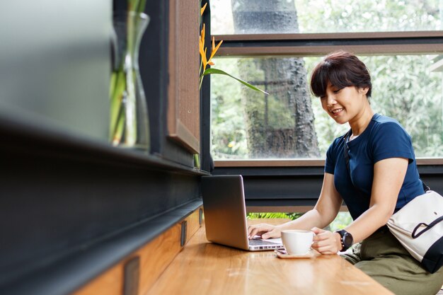 Donna sorridente del viaggiatore che controlla il piano di viaggio di prenotazione sul computer portatile mentre sedendosi al caffè