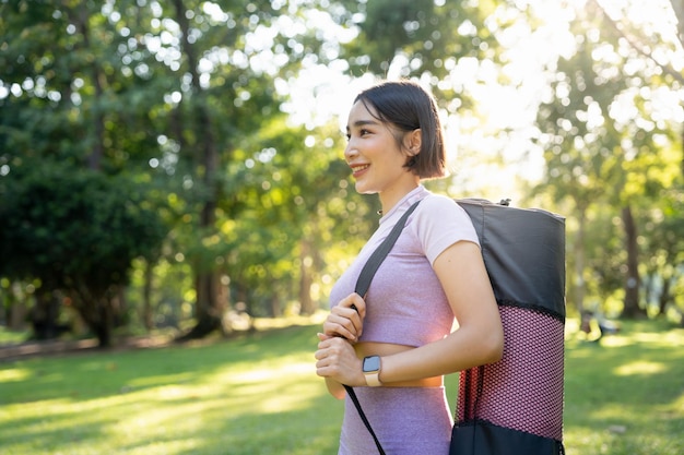 Donna sorridente di forma fisica che sta nel parco del giardino che porta una stuoia di yoga