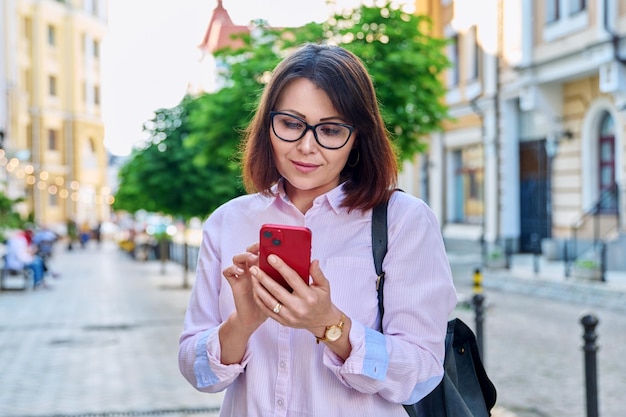 Donna sorridente di mezza età che esamina smartphone sulla strada della città