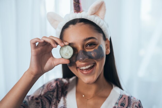 Donna sorridente divertente della stazione termale con il cetriolo della tenuta della maschera facciale fresca