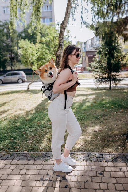 Donna sorridente felice che cammina all'aperto nel parco cittadino con il cane Welsh Corgi Pembroke in uno zaino speciale