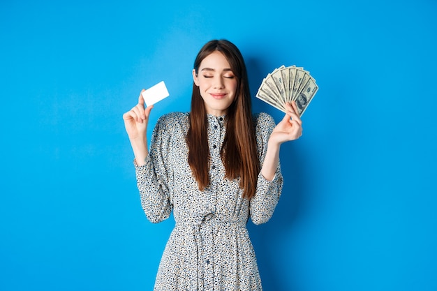 Donna sorridente felice che sembra soddisfatta con gli occhi chiusi, mostrando banconote da un dollaro e carta di credito in plastica, in piedi sognante contro il blu.