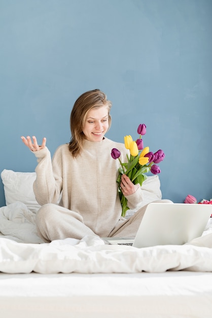 Donna sorridente felice che si siede sul letto che indossa il pigiama, con piacere che gode dei fiori, chiacchierando usando il laptop