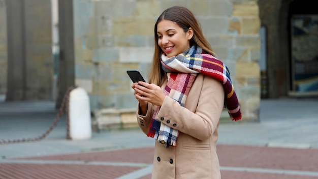 Donna sorridente felice con cappotto e sciarpa utilizzando smart phone all'aperto in una strada cittadina