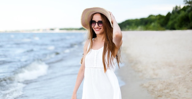 Donna sorridente felice in felicità libera sulla spiaggia dell'oceano in piedi e in posa con cappello e occhiali da sole. Ritratto di una modella femminile in abito bianco d'estate che si gode la natura.