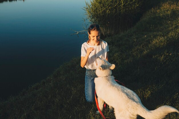 Donna sorridente felice in piedi sulla riva del lago erboso e guardando carino grande cane bianco al guinzaglio rosso nelle vicinanze