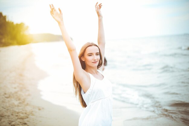 Donna sorridente felice nella beatitudine della felicità libera sulla spiaggia dell'oceano in piedi con le mani alzate. Ritratto di un modello femminile multiculturale in abito estivo bianco che si gode la natura durante le vacanze di viaggio