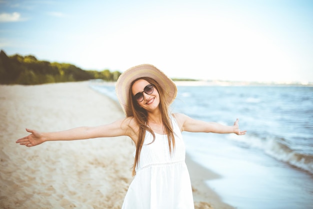 Donna sorridente felice nella beatitudine della felicità libera sulla spiaggia dell'oceano in piedi con un cappello, occhiali da sole e mani aperte. Ritratto di un modello femminile multiculturale in abito estivo bianco che si gode la natura durante il viaggio