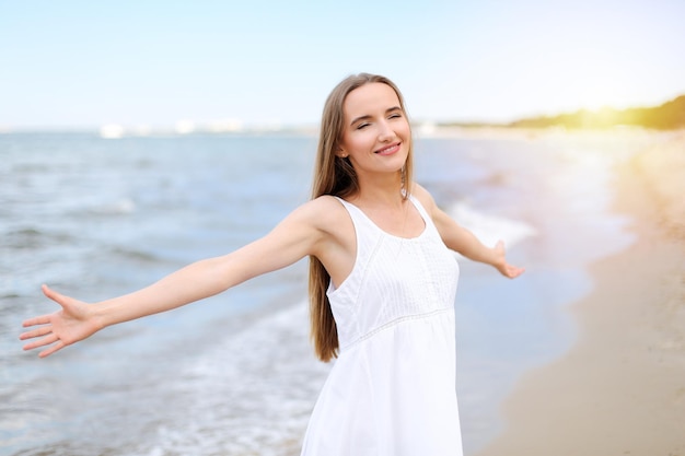Donna sorridente felice nella beatitudine di felicità libera sulla spiaggia dell'oceano in piedi con le mani aperte. Ritratto di un modello femminile multiculturale in abito estivo bianco che si gode la natura durante le vacanze di viaggio
