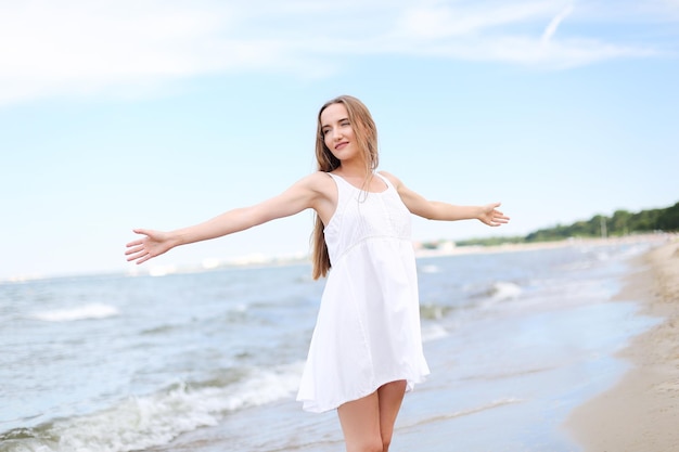 Donna sorridente felice nella felicità libera sulla spiaggia dell'oceano in piedi con le mani aperte. Ritratto di una modella femminile multiculturale in abito estivo bianco che si gode la natura durante le vacanze di viaggio all'aperto