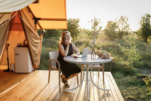 Donna sorridente freelance che parla al telefono mentre lavora in un'accogliente tenda glamping in una giornata di sole Tenda da campeggio di lusso per vacanze estive all'aperto e vacanze Concetto di stile di vita