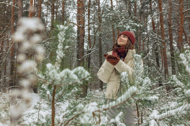 Donna sorridente in capispalla invernali godendo di nevicate nella foresta sempreverde e alzando lo sguardo