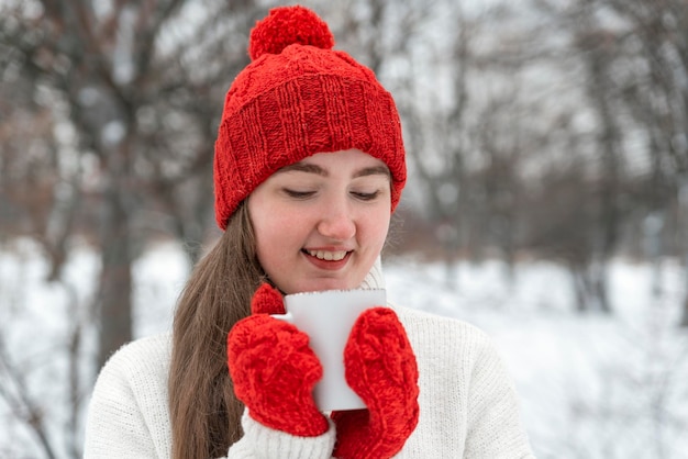 Donna sorridente in cappello di lana rossa e guanti con tazza di bevanda calda in inverno fuori Passeggiata invernale nel parco
