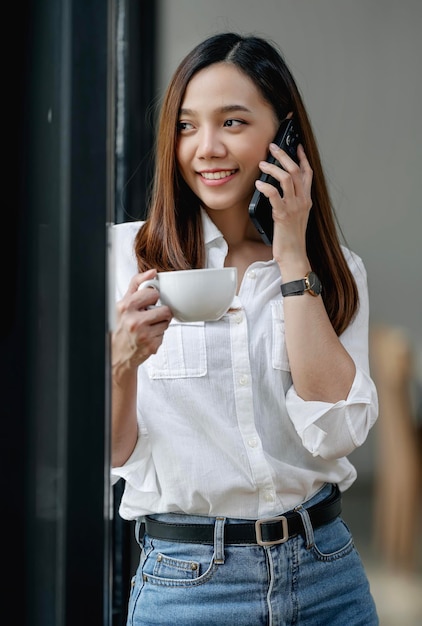 Donna sorridente in casuals azienda tazza di caffè in piedi in ufficio Imprenditrice utilizzando il telefono cellulare e guardando fuori