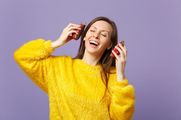 Donna sorridente in maglione di pelliccia che tiene gli occhi chiusi e tiene metà di pitahaya, frutto del drago vicino alle orecchie isolate su sfondo viola pastello. Lo stile di vita vivido delle persone si rilassa il concetto di vacanza. Mock up spazio di copia.