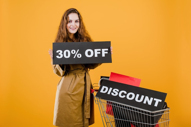 Donna sorridente in trench con il segno di sconto del 30% e sacchetti della spesa variopinti in carrello isolato sopra giallo