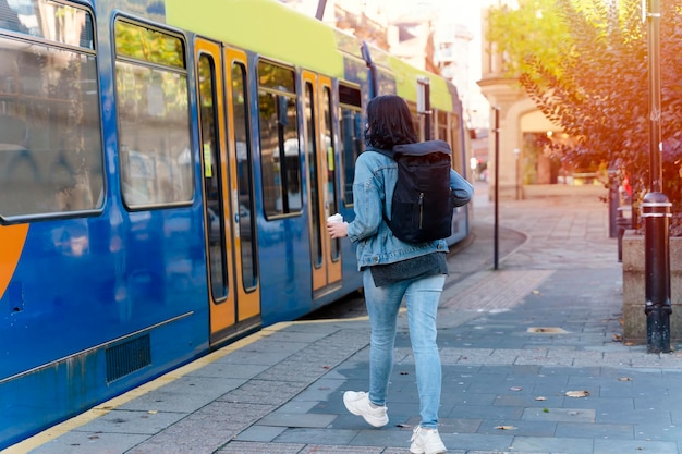 donna sorridente in una giacca di jeans che beve caffè e aspetta un tram alla fermata Foto di stile di vita