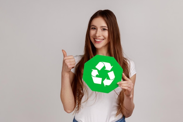 Donna sorridente positiva che mostra i pollici in su come un gesto che tiene il simbolo verde, smista i rifiuti, salva l'ambiente, indossa una maglietta bianca. Studio indoor girato isolato su sfondo grigio.