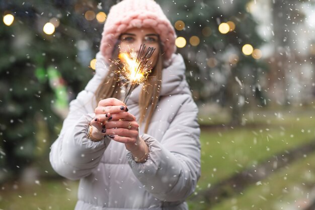 Donna sorridente positiva che si diverte con le stelle filanti vicino all'albero del nuovo anno durante la nevicata