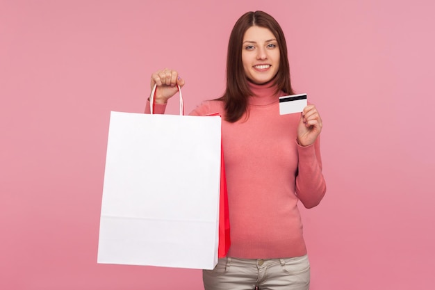 Donna sorridente positiva con capelli castani che tiene e mostra sacchetti di carta e carta di credito, pagamenti contactless, acquisti facili con carta di debito. Studio indoor girato isolato su sfondo rosa