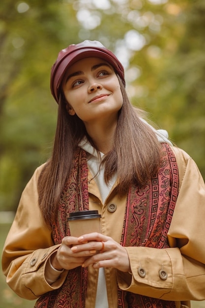 Donna sorridente sognante con una tazza di caffè da asporto in piedi nel parco e godersi il clima autunnale