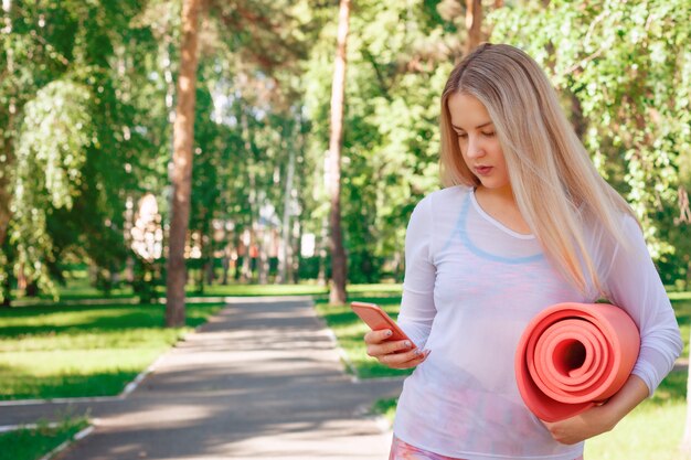 Donna sorridente sveglia che utilizza smartphone e che tiene il materassino yoga nel parco. Piuttosto giovane che trasporta una stuoia di yoga e mandare SMS sul suo telefono cellulare all'aperto