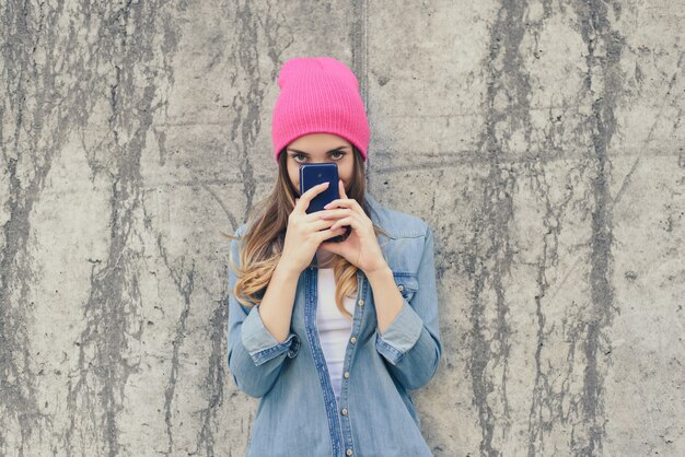 Donna sospettosa o timida in abiti casual e cappello rosa che nasconde il viso dietro lo smartphone, legge le informazioni del settore. La donna è in piedi sulla strada contro la superficie grigia