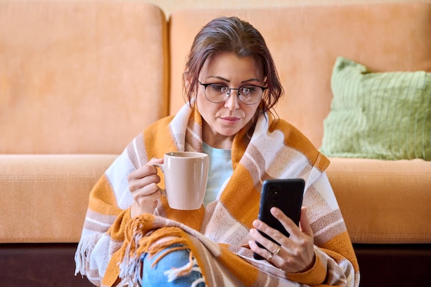 Donna sotto una calda coperta di lana con una tazza di tè caldo utilizzando uno smartphone