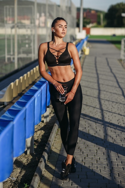 Donna sportiva attraente con le cuffie e la bottiglia senza fili di acqua che ascolta la musica mentre si prepara su un campo sportivo all'aperto.