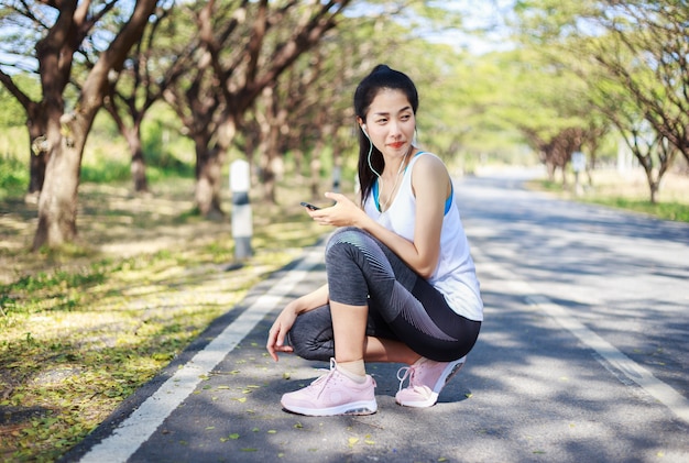 donna sportiva che si siede sulla strada nel parco e utilizzando smartphone con auricolare per ascoltare musica