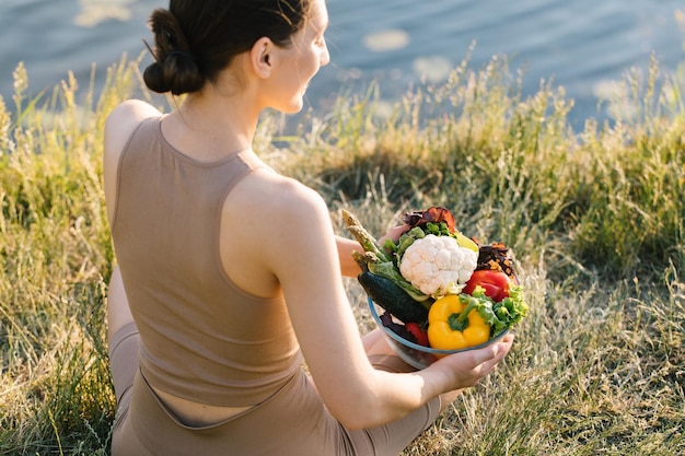 Donna sportiva che tiene un piatto di verdure fresche all'aperto vicino al fiume vista posteriore