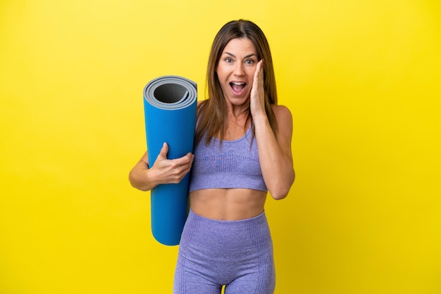 Donna sportiva che va a lezioni di yoga mentre tiene un tappetino isolato su sfondo non giallo con espressione facciale sorpresa e scioccata