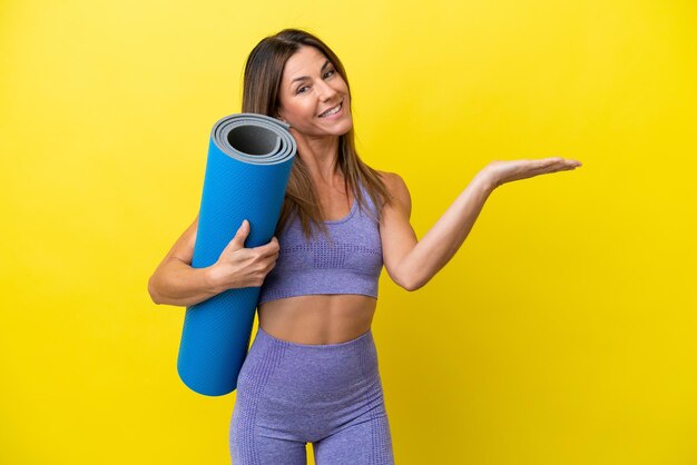 Donna sportiva che va a lezioni di yoga mentre tiene un tappetino isolato su uno sfondo non giallo che estende le mani di lato per invitare a venire