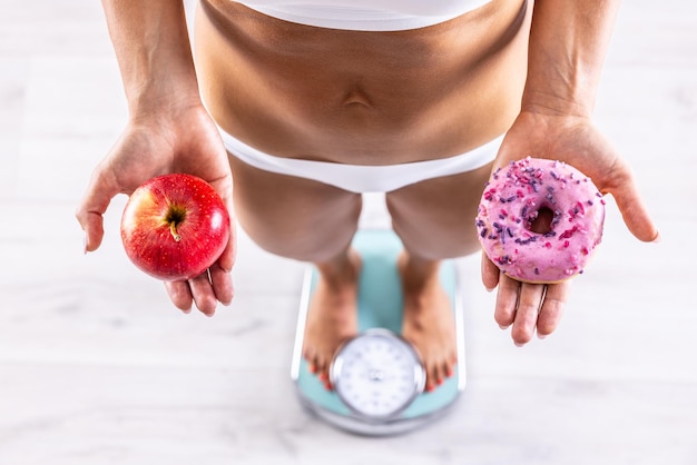 Donna sportiva con un corpo perfetto che misura il peso sulla bilancia e tiene in mano una ciambella e una mela rossa Concetto di stile di vita sano o malsano