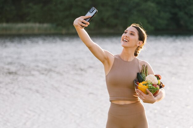 Donna sportiva felice che prende selfie con un piatto di verdure fresche all'aperto