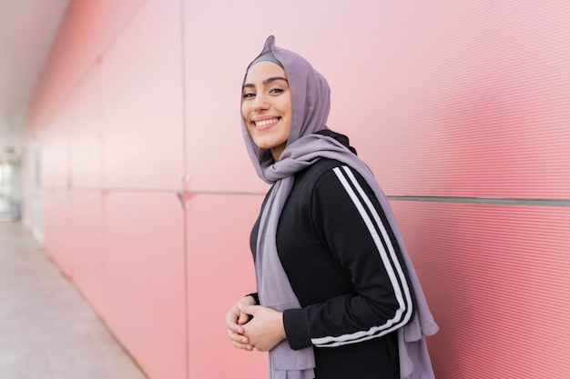 Donna sportiva in hijab pronta a correre su un muro rosso guardando di lato con un sorriso sul viso