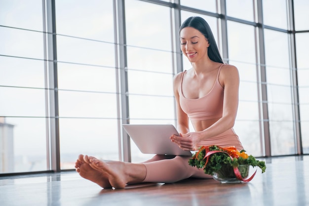 Donna sportiva seduta sul tavolo con laptop e cibo sano.