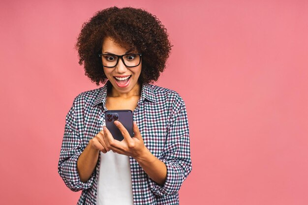 Donna studentessa stupita e scioccata afroamericana che utilizza lo smartphone in piedi sopra isolato su sfondo rosa Utilizzo del telefono cellulare