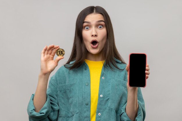 Donna stupita che tiene bitcoin d'oro e smartphone con schermo vuoto per pubblicità o promozione