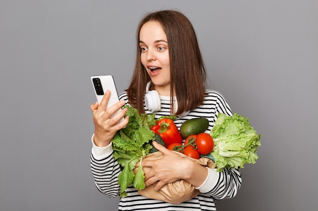Donna stupita che tiene le verdure isolate su sfondo grigio tenendo il telefono cellulare utilizzando internet alla ricerca di informazioni su un'alimentazione sana