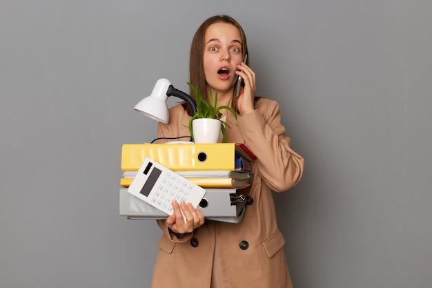 Donna stupita e stupita che indossa un'elegante giacca beige che tiene roba da ufficio in posa isolata su sfondo grigio che parla sullo smartphone sentendo qualcosa di scioccato ultime notizie sul suo licenziamento