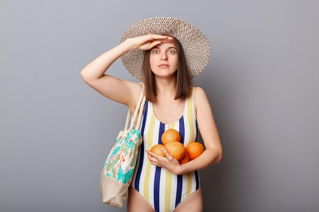 Donna stupita sorpresa scioccata che indossa costume da bagno a righe e cappello di paglia isolato sfondo grigio in piedi con arance fresche tiene la mano vicino alla fronte guardando lontano