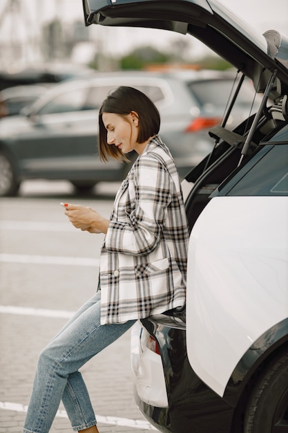 Donna sulla stazione di ricarica per auto elettriche durante il giorno. Donna seduta sul bagagliaio aperto della sua auto elettrica e usa un telefono