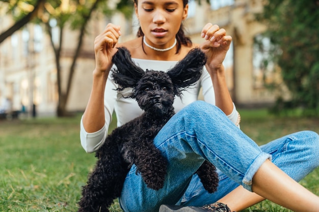 Donna sveglia che gioca con un piccolo cane nero divertente che si siede sull'erba nel parco