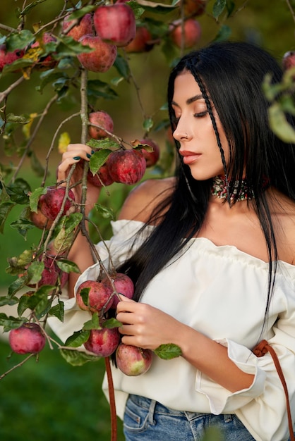 Donna triste dai capelli scuri con la camicia bianca senza spalle che tiene i rami degli alberi carichi di mele rosse vicino