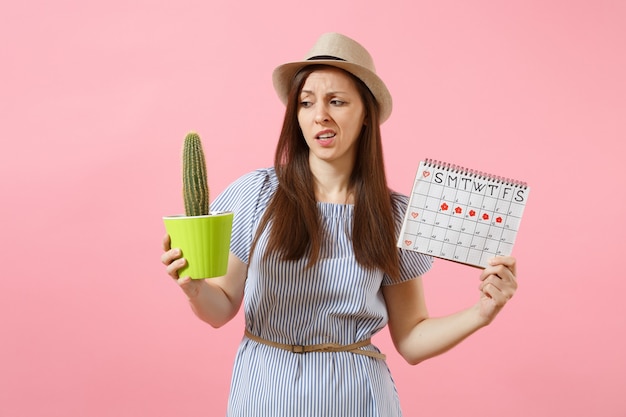 Donna triste di malattia in vestito blu che tiene cactus verde, calendario dei periodi per controllare i giorni delle mestruazioni isolati su sfondo rosa. Medico, sanitario, ginecologico, concetto di dolore alla pancia. Copia spazio.