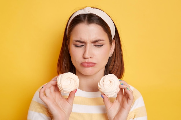 Donna triste sconvolta che indossa maglietta e fascia per capelli in posa isolata su sfondo giallo essendo a dieta ma vuole mangiare marshmallow zuccherati guardando il dessert con le labbra imbronciate ed esprimendo dolore