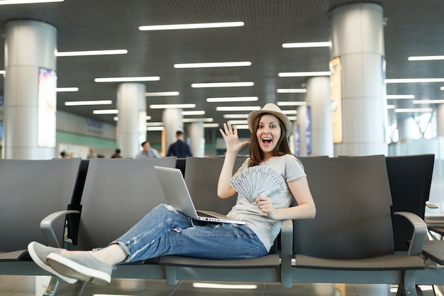 Donna turistica sorpresa che lavora al computer portatile, tiene in mano un pacco di dollari in contanti, allarga le mani in attesa nella hall dell'aeroporto